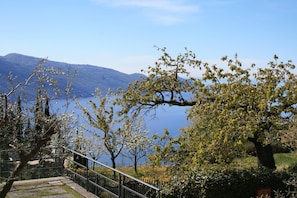 A scenic view of a mountainous landscape with a lake, a pathway with a railing, and trees in the foreground. - Appartamento Anna 701 (Tignale)