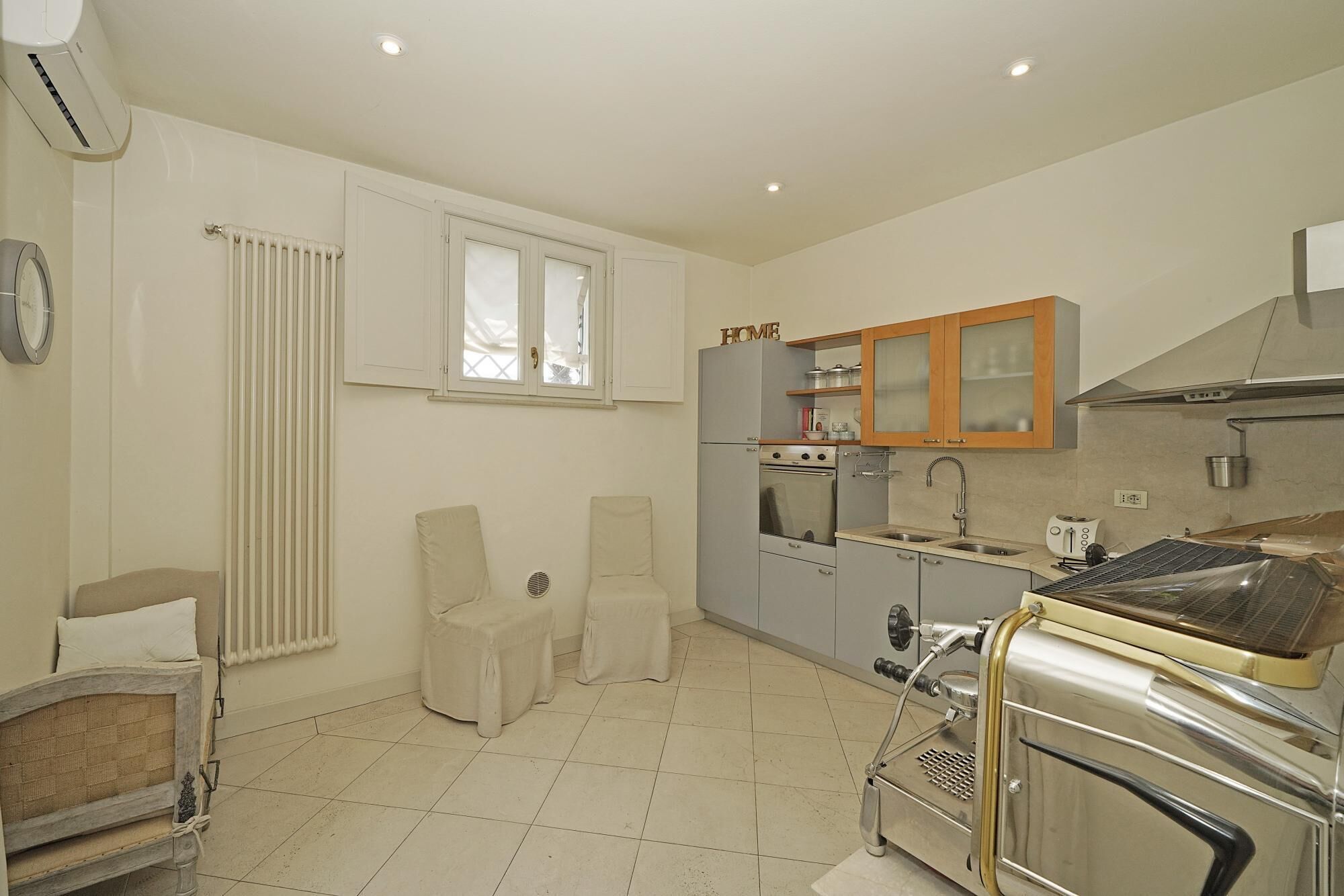 Cocina pequeña con refrigerador, estufa y gabinetes de madera. Se aprecia una ventana con persianas, un radiador y un reloj en la pared.