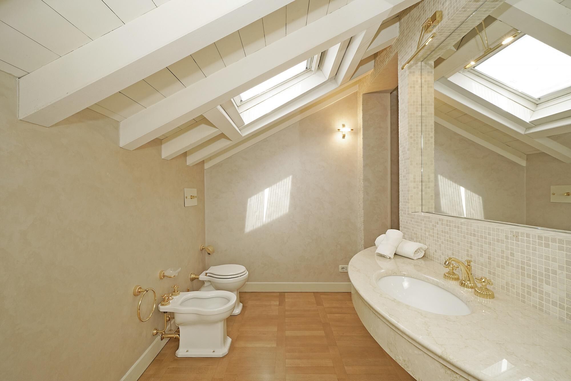 Un baño con tragaluz, un espejo grande y un lavamanos blanco con detalles dorados.