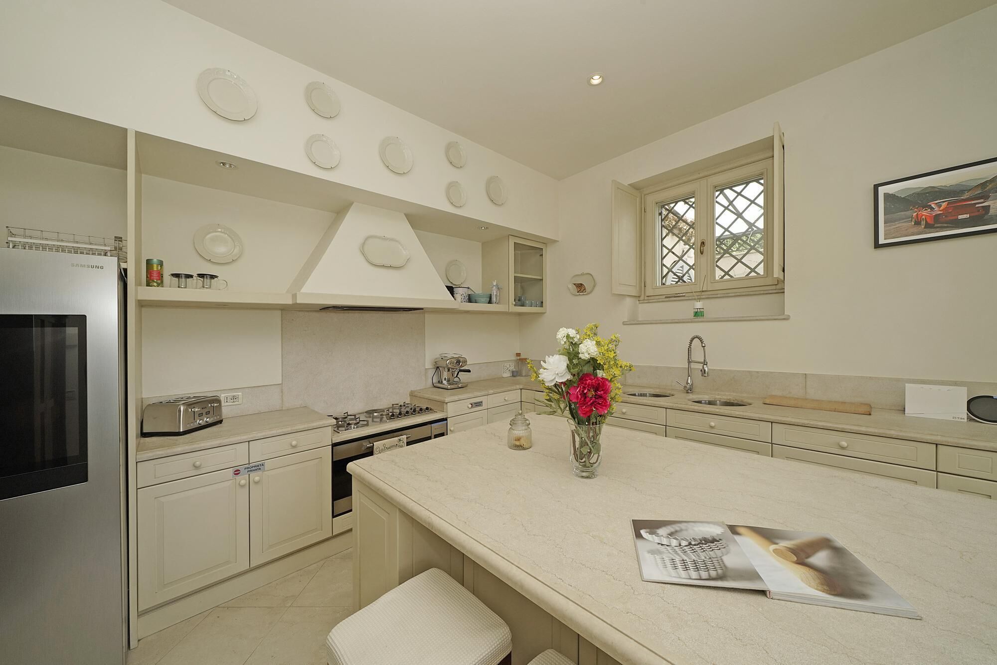 A modern kitchen with a stainless steel refrigerator, a gas stove, and a sink. There is a vase with flowers on the island, and a framed picture on the wall.
