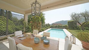 A poolside dining area with a wooden table, white chairs, and a view of a swimming pool and hills in the background. - Villa San Carlo in Ro (Roe Volciano)