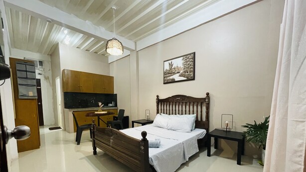 A bedroom with a wooden bed, a nightstand, a desk, a chair, a potted plant, and a kitchen area in the background. - Sto. Nino Residences (Lucena)