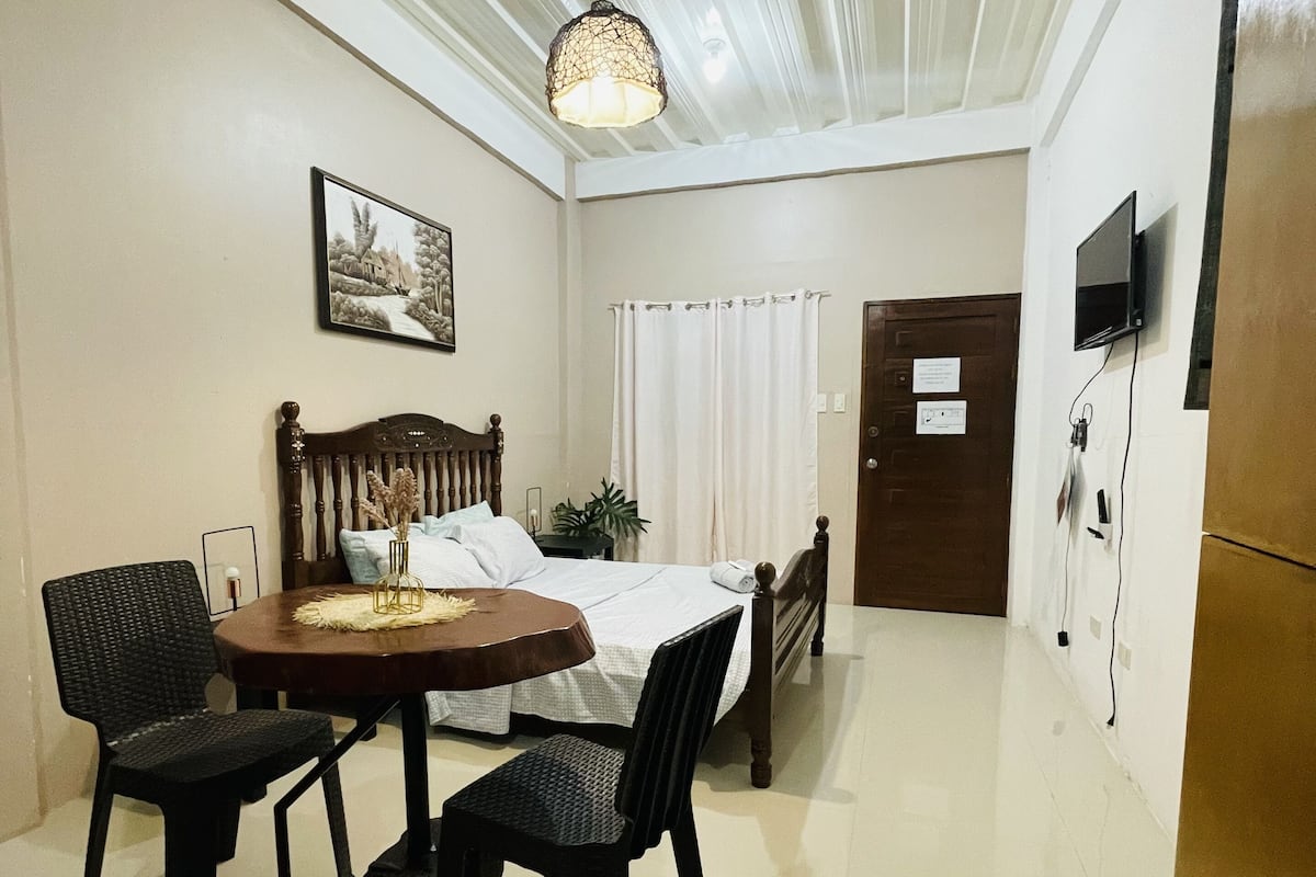 A hotel room with a bed, a dining table with chairs, a ceiling light, a television, and a door.
