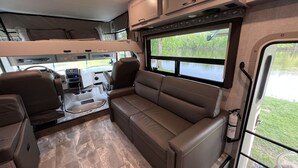 A compact RV interior with a kitchenette, a seating area, and a window offering a view of a lake.