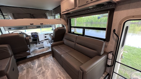 A compact RV interior with a kitchenette, a seating area, and a window offering a view of a lake. - Luxury RV with Bunkbeds Stationed on serene Grand River with private golf course (Grand Rapids)
