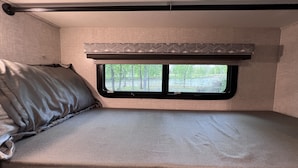 A compact RV interior with a bed, a window with a view of trees, and a patterned headboard.