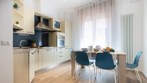 A modern kitchen with white cabinets, a stainless steel range hood, and a built-in oven. There is a dining area with a wooden table and blue chairs. - Nida 18 in Cattolica (Cattolica)