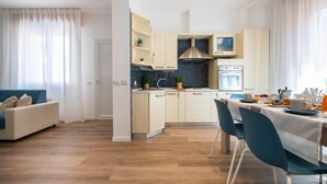 A modern kitchen with white cabinets, a stainless steel range hood, and a built-in oven. There is a dining area with a table and chairs, and a sofa in the living room. - Nida 18 in Cattolica (Cattolica)