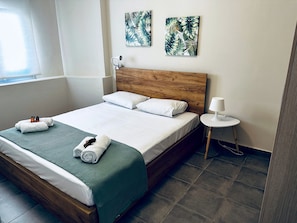 Ein ordentlich bezogenes Bett mit weißen Leinen, einer grünen Decke und aufgerollten Handtüchern. Zwei an der Wand befestigte Kunstwerke zeigen tropische Blätter. Ein kleiner weißer Tisch mit einer Lampe steht neben dem Bett.