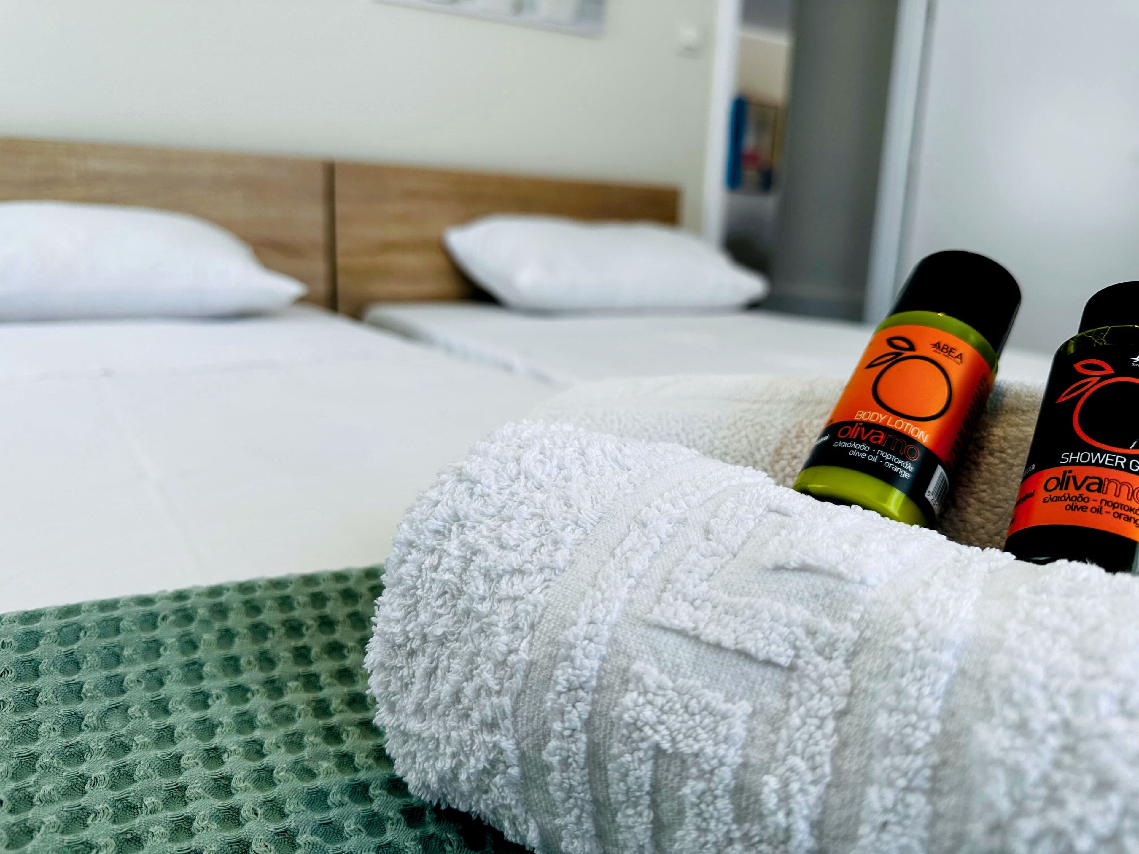 A hotel room with a neatly made bed, two bottles of body lotion, and a rolled-up towel on a green mat.