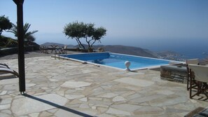 Una piscina infinita con un patio de piedra, mobiliario exterior y vistas al mar y las montañas.