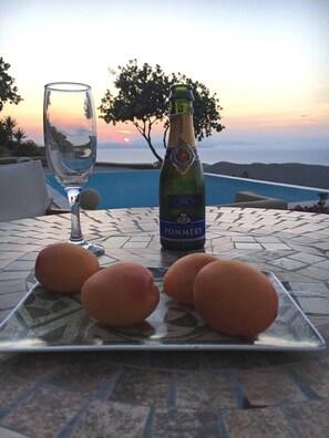 Una mesa con una botella de champán Pommery, una copa y tres huevos, sobre una mesa con baldosas y vistas a una piscina y al atardecer.
