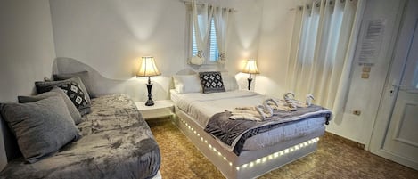 A bedroom with a bed, a sofa, two bedside tables with lamps, a window with curtains, and a door with a sign.