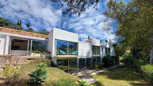 A modern house with a glass facade, a stone foundation, and a garden with various plants. - Villa zu Vermieten in Porto del Almendros, Los Arqueros, Benahavis (Benahavís)
