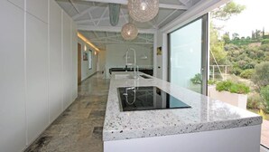 A modern kitchen with a central island, large windows, and a view of a hilly landscape. - Villa zu Vermieten in Porto del Almendros, Los Arqueros, Benahavis (Benahavís)