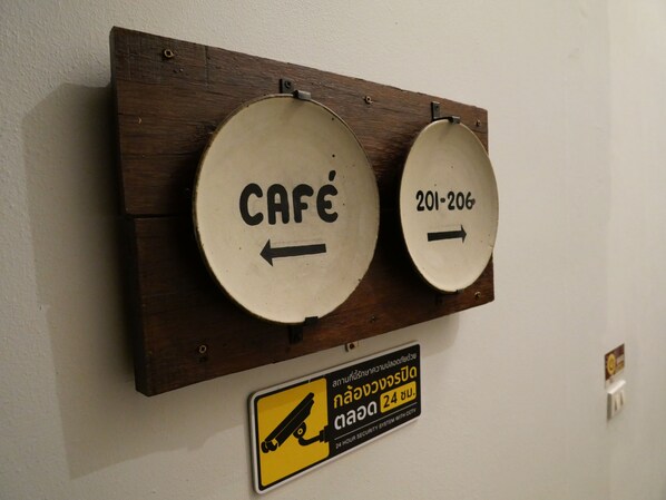 A wall-mounted sign with two circular labels, one labeled 'CAFÉ' with an arrow pointing left, and the other labeled '201-206' with an arrow pointing right. Below the labels, there is a yellow sign with a security camera symbol and text indicating a 24-hour security system.