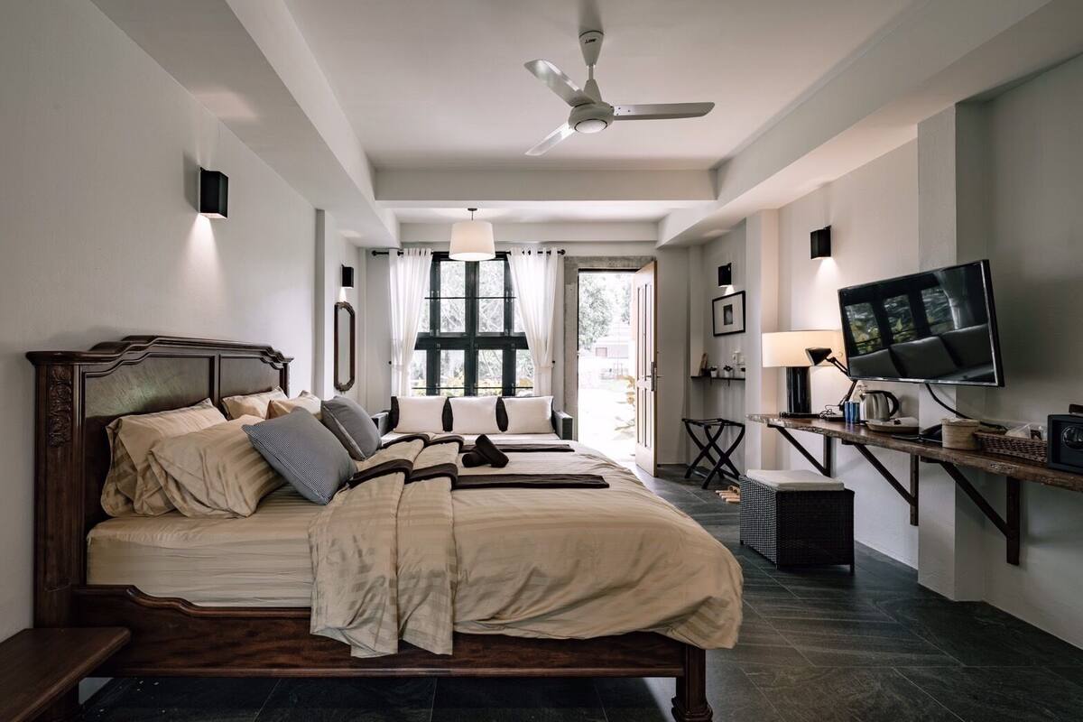 A bedroom with a large bed, a desk with a TV, a ceiling fan, and a window with curtains.