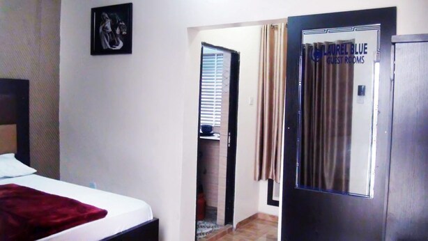 A hotel room with a bed, a door labeled "LAUREL BLUE GUEST ROOMS," and a small window with blinds. - Laurel Blue Suites (Port Harcourt)
