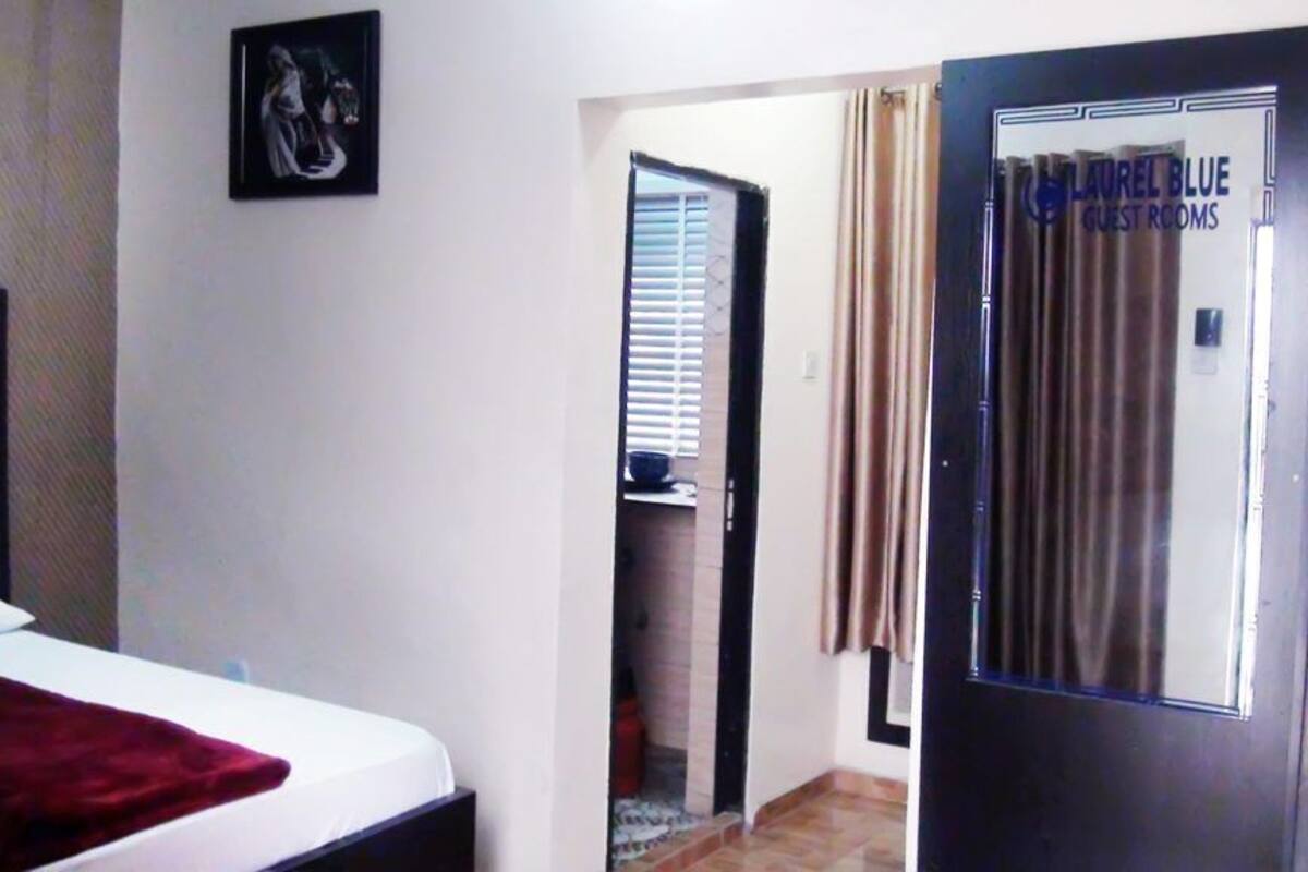 A hotel room with a bed, a door labeled "LAUREL BLUE GUEST ROOMS," and a small window with blinds.