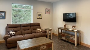 Een woonkamer met een bruine bank, een salontafel, een televisie aan de muur en ingelijste foto's.