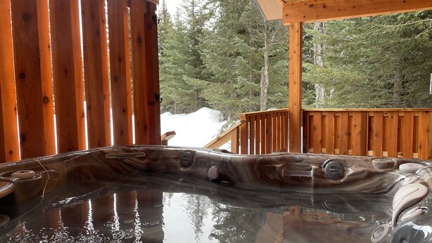Een hottub met een houten buitenkant, omgeven door een houten terras en een besneeerd landschap met bomen op de achtergrond.
