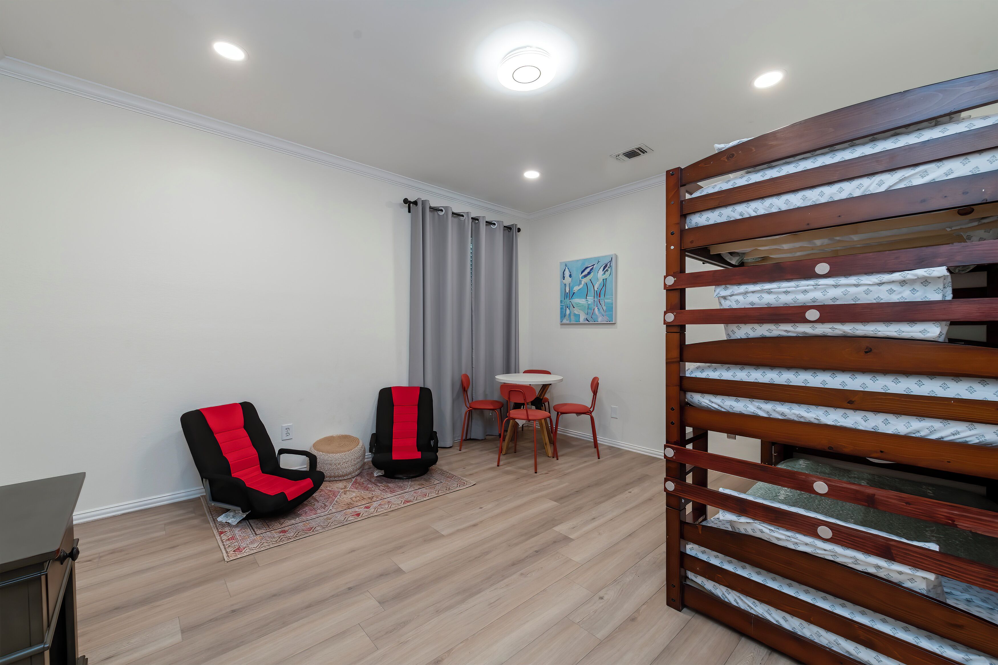 A room with a wooden bed frame, a red and black chair, a small table with chairs, and a grey curtain.