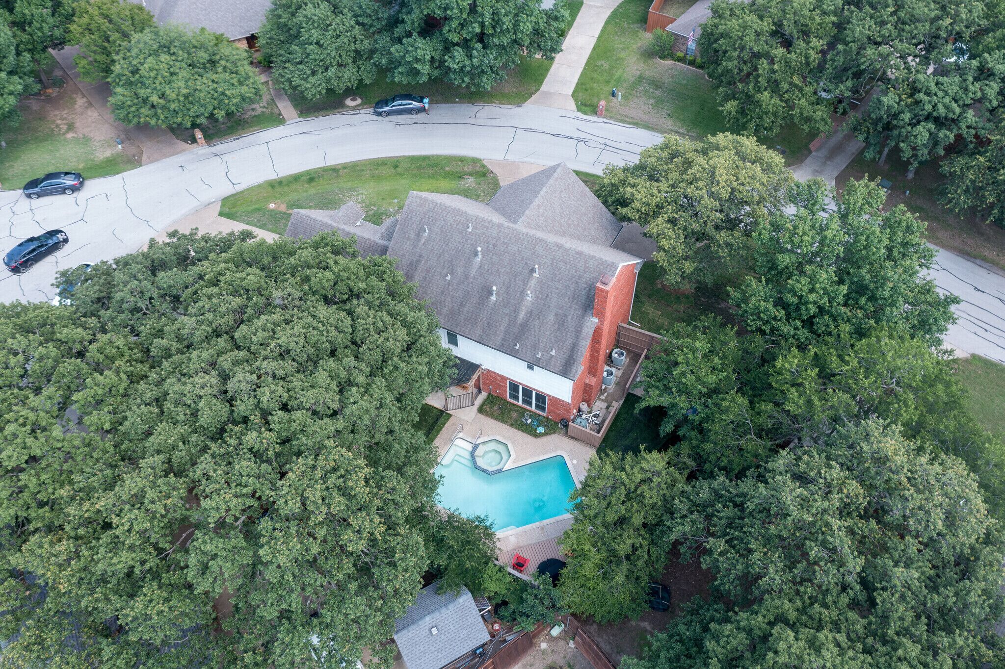 An aerial view of a house with a swimming pool surrounded by dense trees, a driveway with cars, and a red brick building.