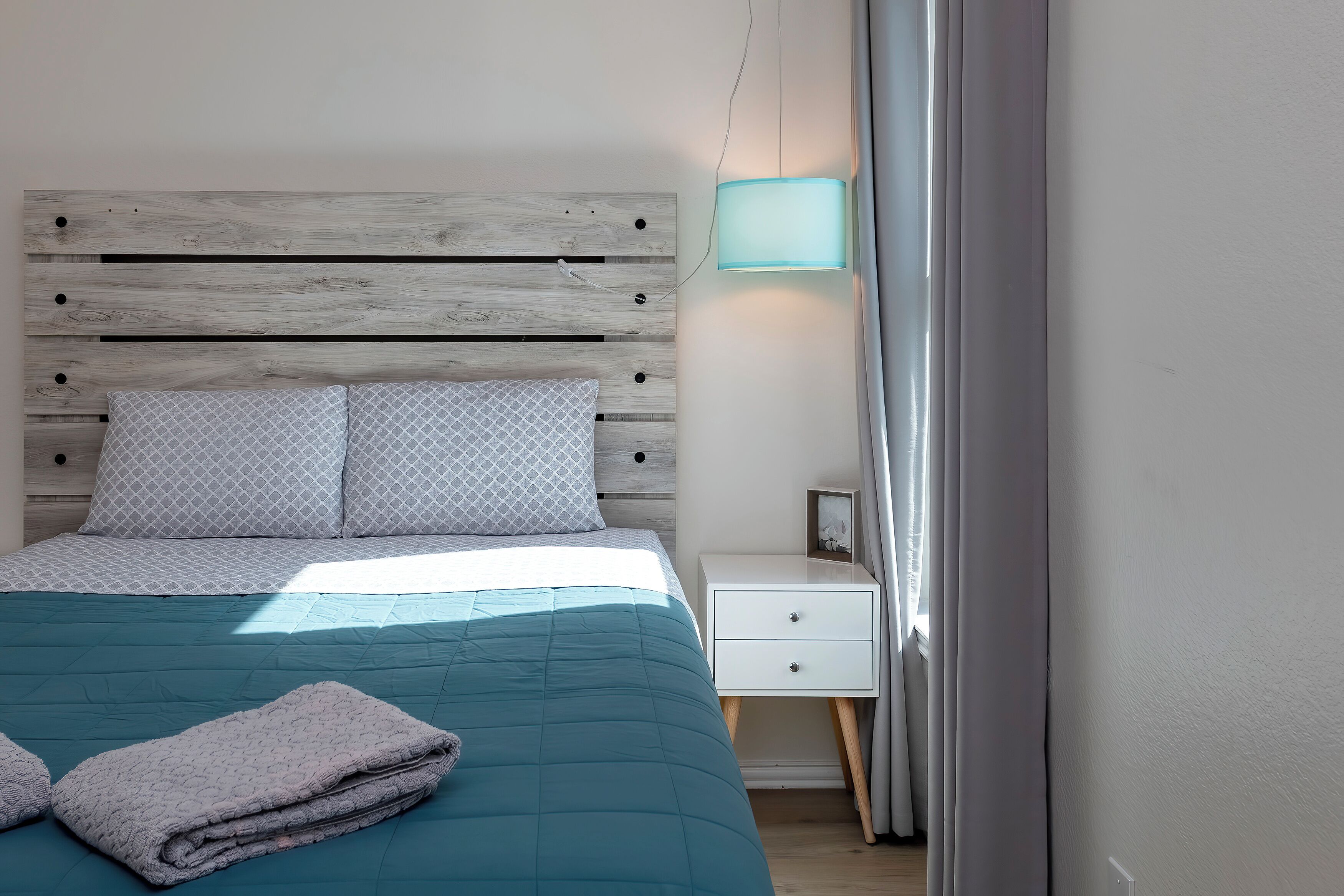 A bedroom with a wooden headboard, a teal bedspread, two pillows, a bedside table with a lamp, and a nightstand with a picture frame.