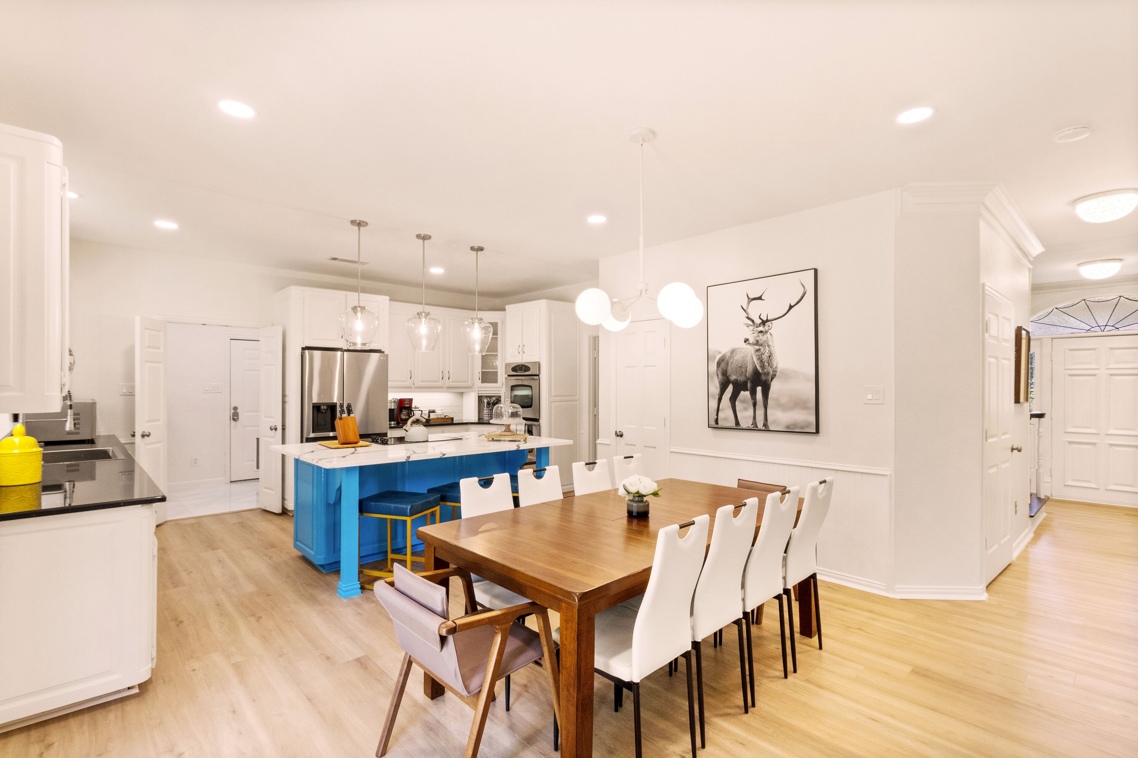 Een moderne keuken met witte kasten, een blauw kookeiland en een eethoek met een houten tafel en witte stoelen.