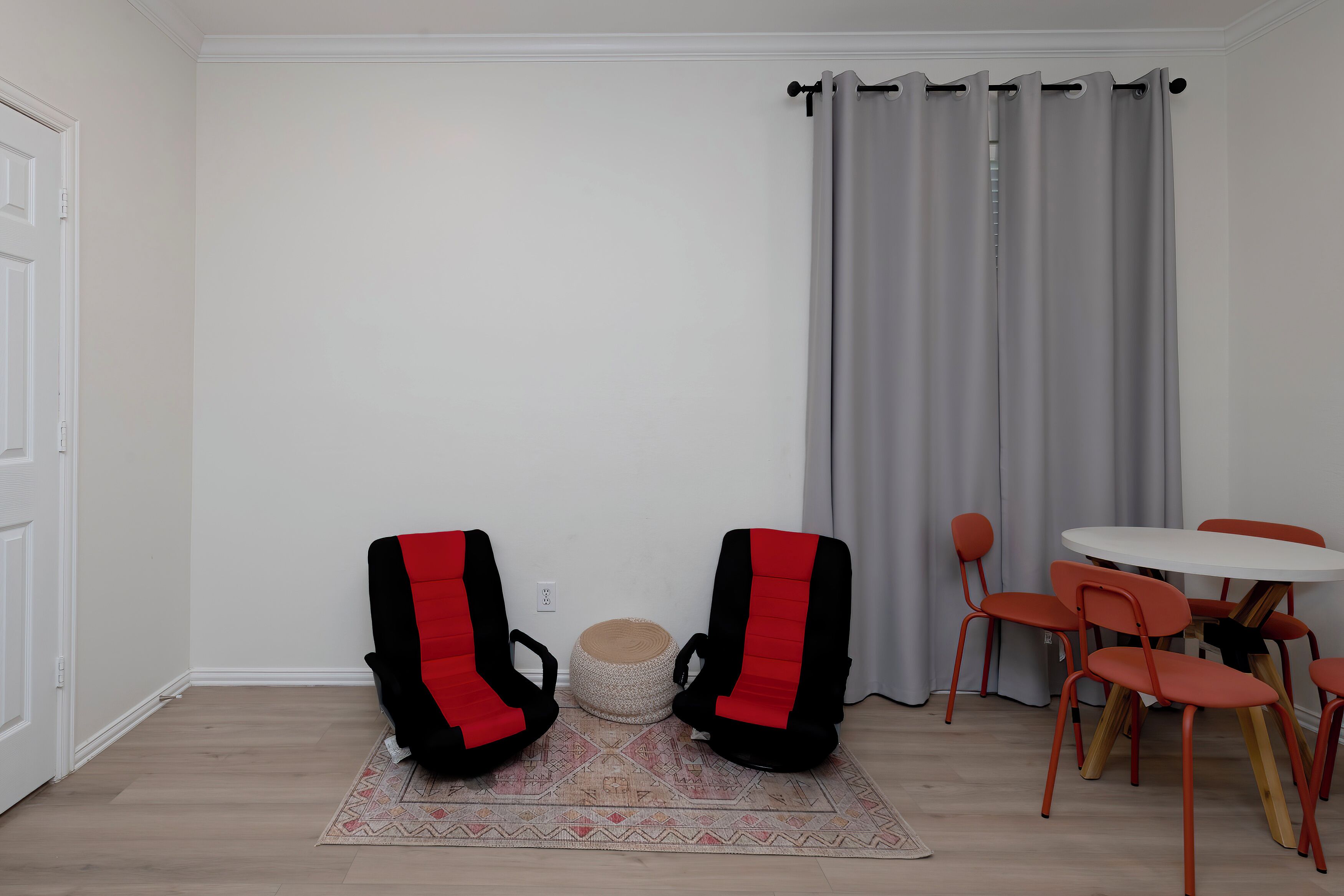 Ett rum med två röda och svarta stolar, ett runt bord med sex orangefärgade stolar, en flätad pall och en grå gardin.