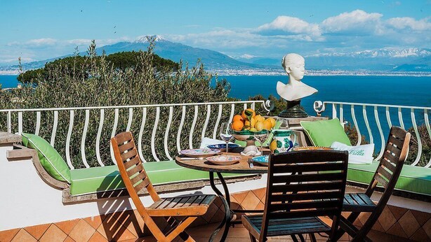 Eine Terrasse mit Esstisch, einer Büstenstatue und Blick auf Meer und Berge.