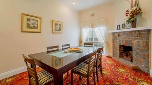 A dining area with a wooden table, chairs, and a red patterned carpet. There is a fireplace with a brick surround and a window with sheer curtains. - Cambridge House, Bendigo (Bendigo)