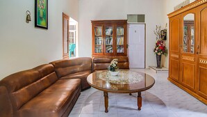 Un salon avec un canapé en cuir marron, une table basse en verre ornée d’un chemin de table à motifs floraux, un meuble en bois rempli de livres et une porte blanche.