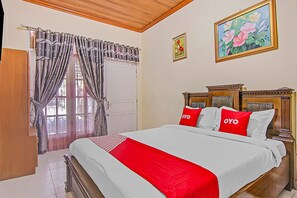 A bedroom with a large bed, two pillows with 'OYO' branding, a wooden headboard, a painting on the wall, and a window with curtains.