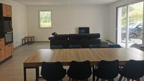 Una cocina moderna con electrodomésticos integrados, una zona de comedor con mesa y sillas de madera, un sofá negro y un televisor sobre un mueble.