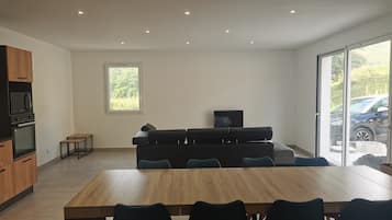 Een moderne keuken met inbouwapparatuur, een eethoek met een houten tafel en stoelen, een zwarte bank en een tv op een standaard.