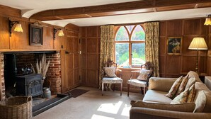A cozy living room with a fireplace, wooden walls, and a large window with curtains. There are two chairs and a sofa with floral cushions. - The Cottage Guest House (Bishop's Stortford)