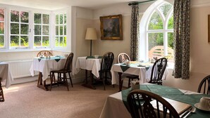 A dining room with four tables, each set with white tablecloths and wooden chairs. There is a painting on the wall and a window with patterned curtains. - The Cottage Guest House (Bishop's Stortford)