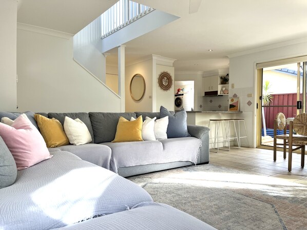 A modern living room with a grey sectional sofa, a staircase, and a view of the kitchen and outdoor area.