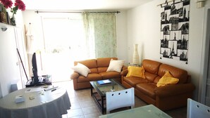 Een woonkamer met een bruine leren bank, een glazen salontafel, een eettafel met een tafelkleed met patroon, en een wand met reiskunst.