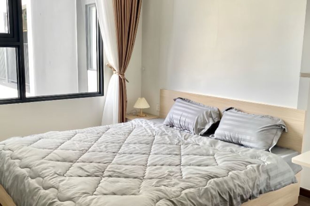 Una cama bien hecha con un edredón de color claro y dos almohadas, una lámpara de noche y una ventana con cortinas.