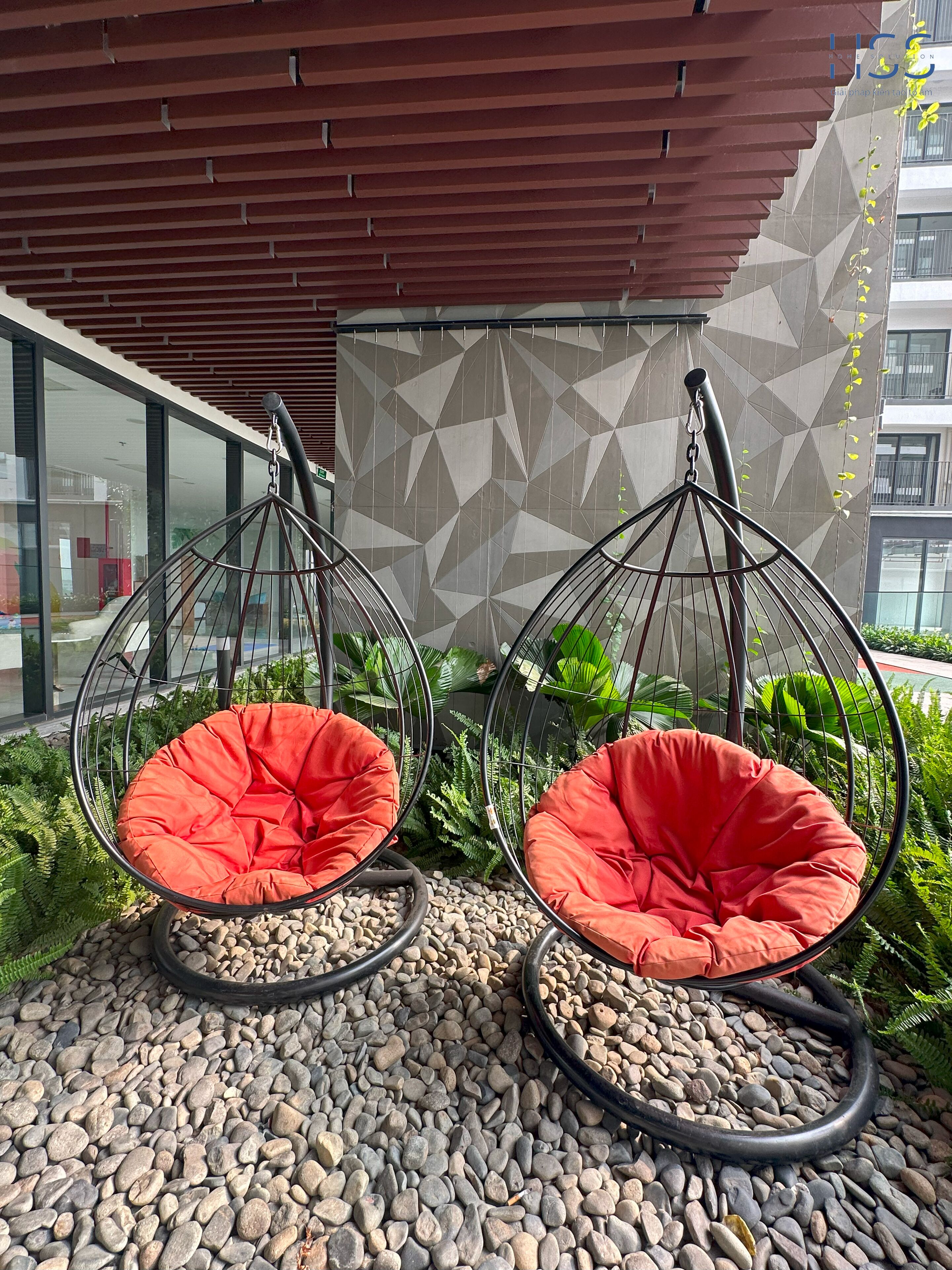 Two red hanging chairs with metal frames, set on a pebbled ground, with a geometric patterned wall and a building in the background.
