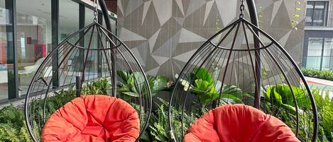 Two red hanging chairs with metal frames, set on a pebbled ground, with a geometric patterned wall and a building in the background.