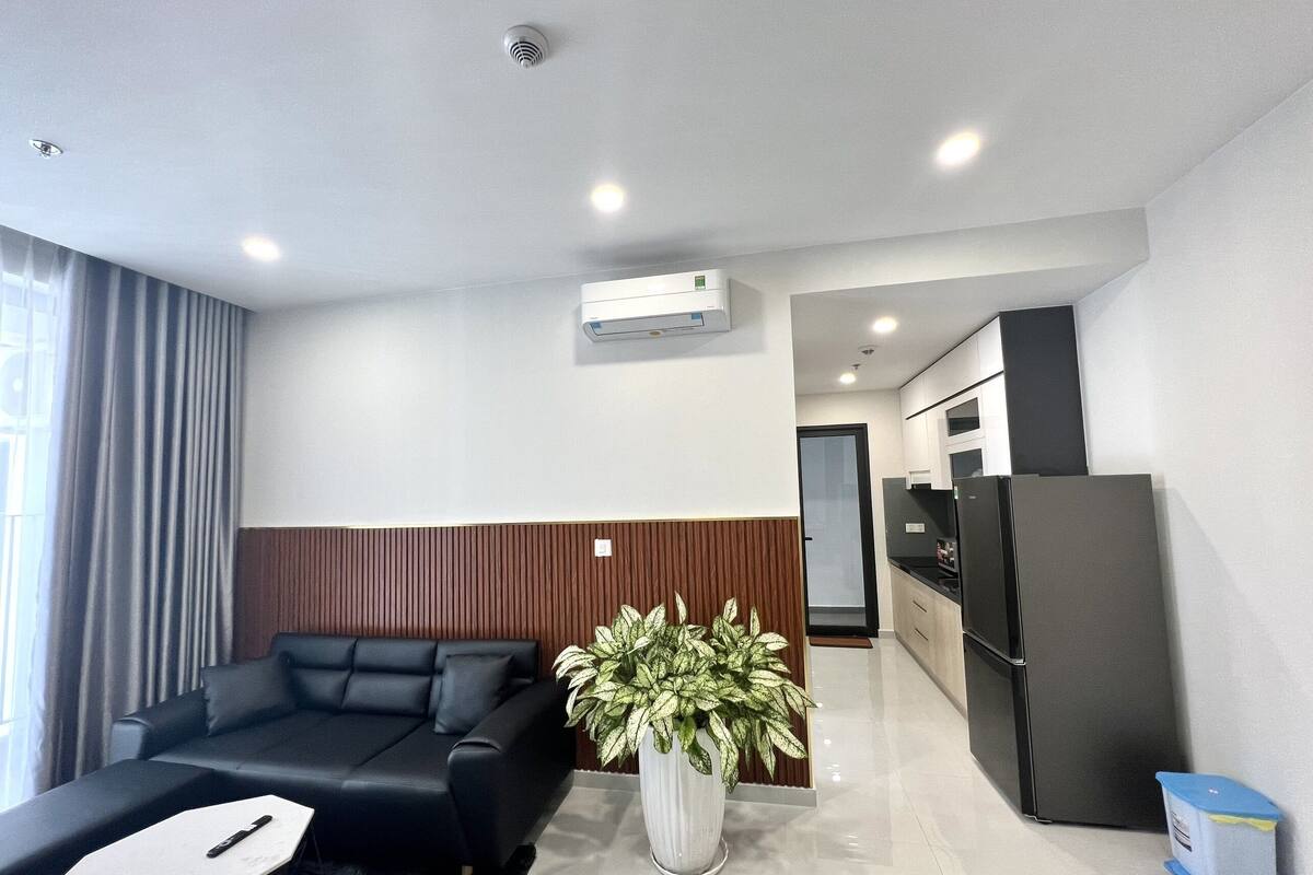 A modern living room with a black sofa, a white coffee table, and a potted plant. There is a kitchen area with a black refrigerator and cabinets.