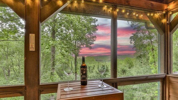 A wooden cabin with a table and two wine glasses, a bottle of wine, and a view of a forest through large windows. - North Georgia Hideaway | Ellijay, GA (Ellijay)