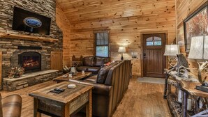 A log cabin interior with a stone fireplace, leather seating, and a mounted television. - North Georgia Hideaway | Ellijay, GA (Ellijay)