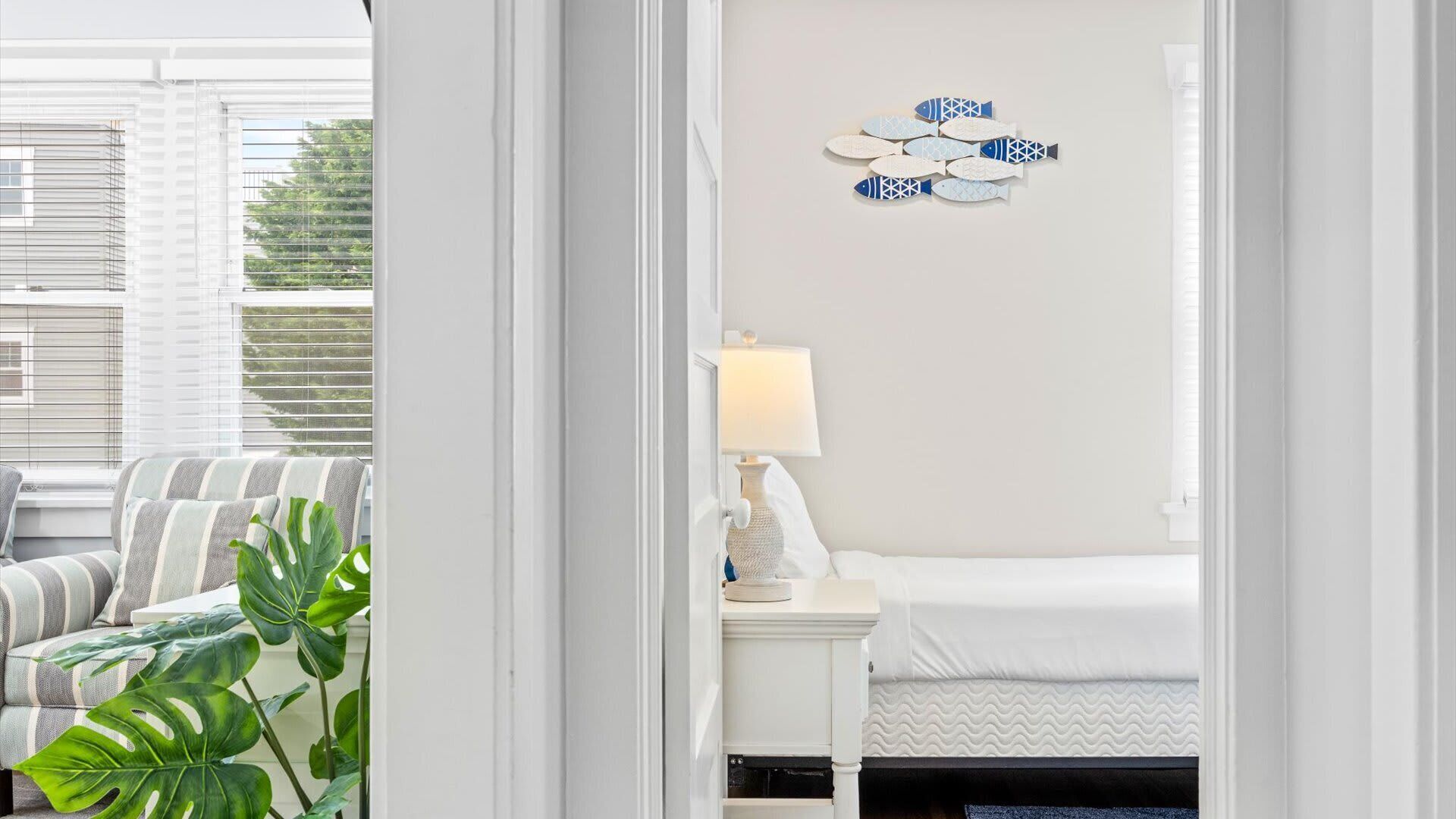 Une chambre d’hôtel avec un lit, une table de chevet avec une lampe, une décoration murale représentant des poissons et une vue sur l’extérieur par une fenêtre équipée de stores.