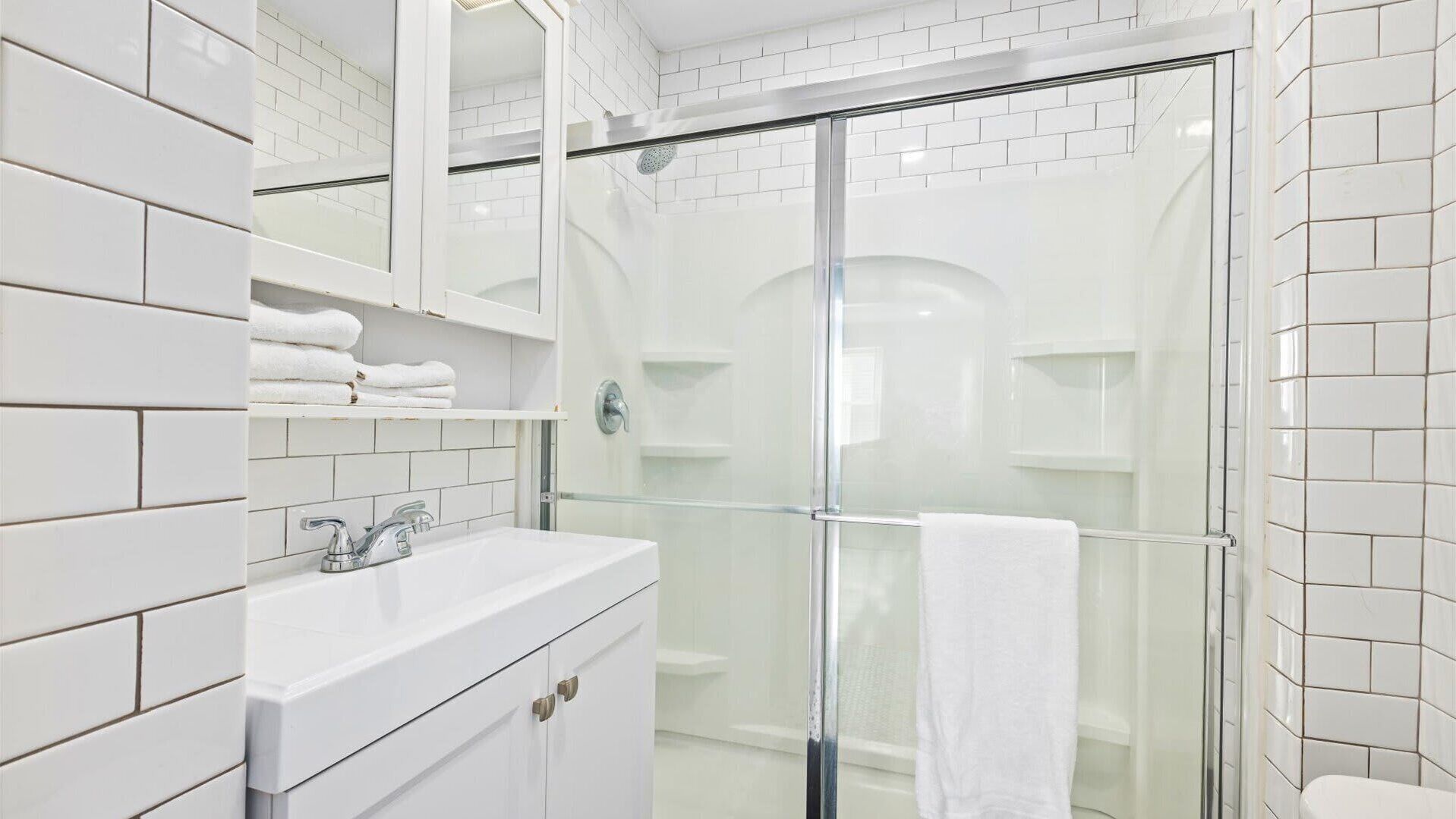Un baño moderno con azulejos blancos, cabina de ducha con mampara de vidrio y tocador con espejo y toallero.