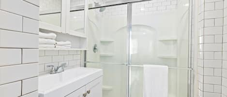 Un baño moderno con azulejos blancos, cabina de ducha con mampara de vidrio y tocador con espejo y toallero.
