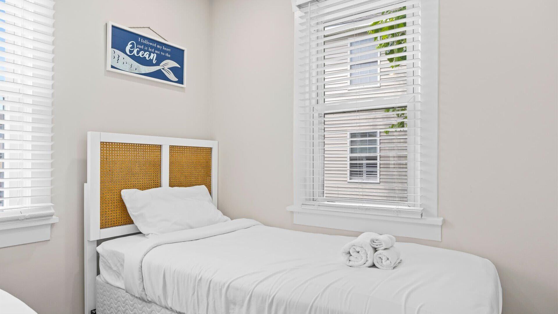 A bedroom with a bed, white bedding, a headboard with a woven panel, and a window with white blinds.